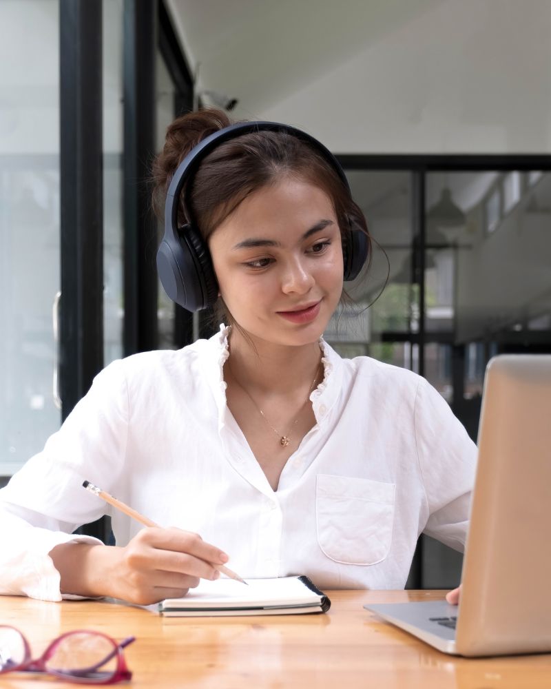 Girl with white shirt, a pen, paper and head set in front of a laptop