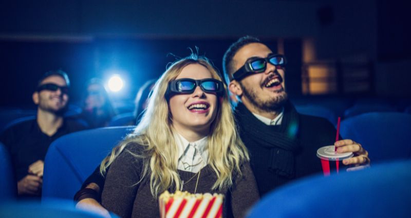 Man and woman smiling with popcorn and 3D glasses in movie theatre